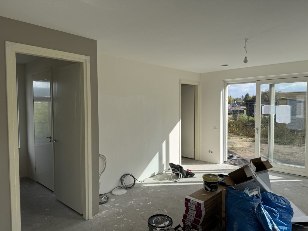 Woonkamer van een nieuwbouwwoning tijdens de afwerking, met verfemmers en dozen op de kale vloer