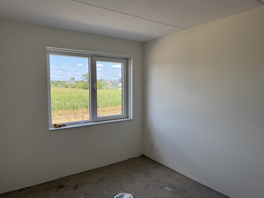Slaapkamer in een nieuwbouwwoning met gladde, lichte wanden en uitzicht op een akker